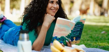 Smiling woman reading a book at a picnic in a sunny park setting.