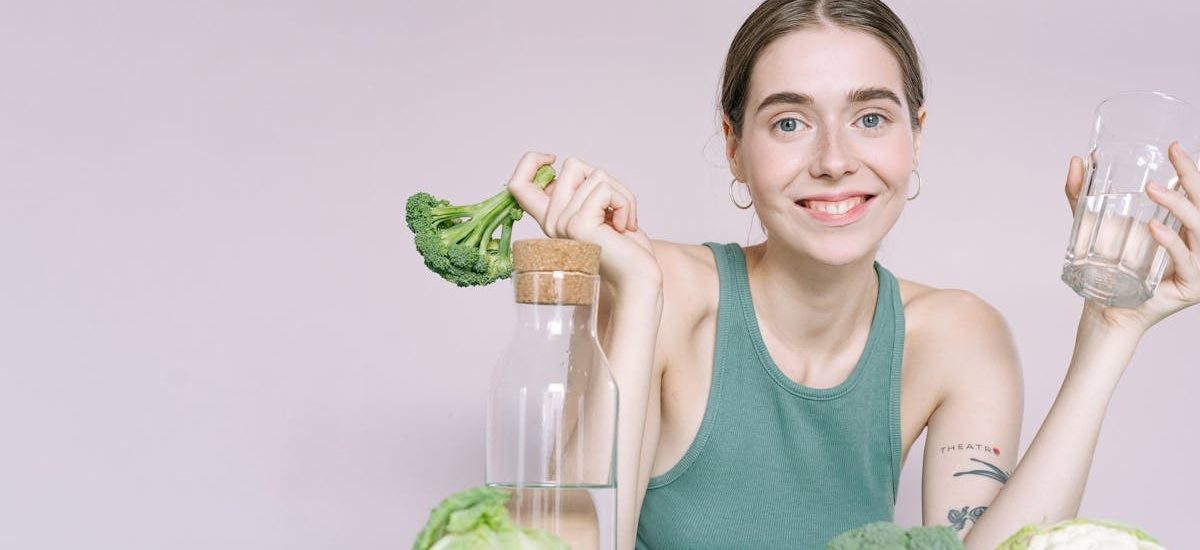 Woman in tank top smiling with broccoli, cabbage, and glass of water on table.