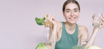 Woman in tank top smiling with broccoli, cabbage, and glass of water on table.