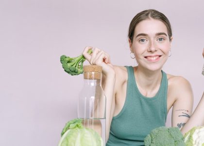 Woman in tank top smiling with broccoli, cabbage, and glass of water on table.